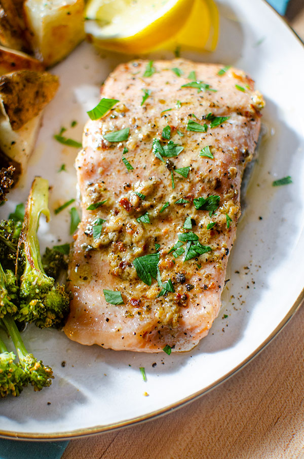 Closeup of trout fillet on a plate with roasted potatoes, broccoli and lemon wedges.