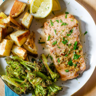 Baked trout fillet on a plate with roasted potatoes, broccoli and lemon wedges.
