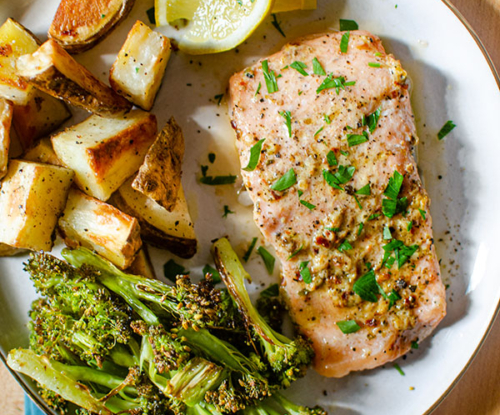 Baked trout fillet on a plate with roasted potatoes, broccoli and lemon wedges.