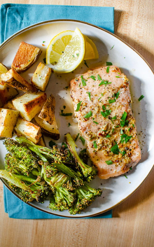 Baked trout fillet on a plate with roasted potatoes, broccoli and lemon wedges.