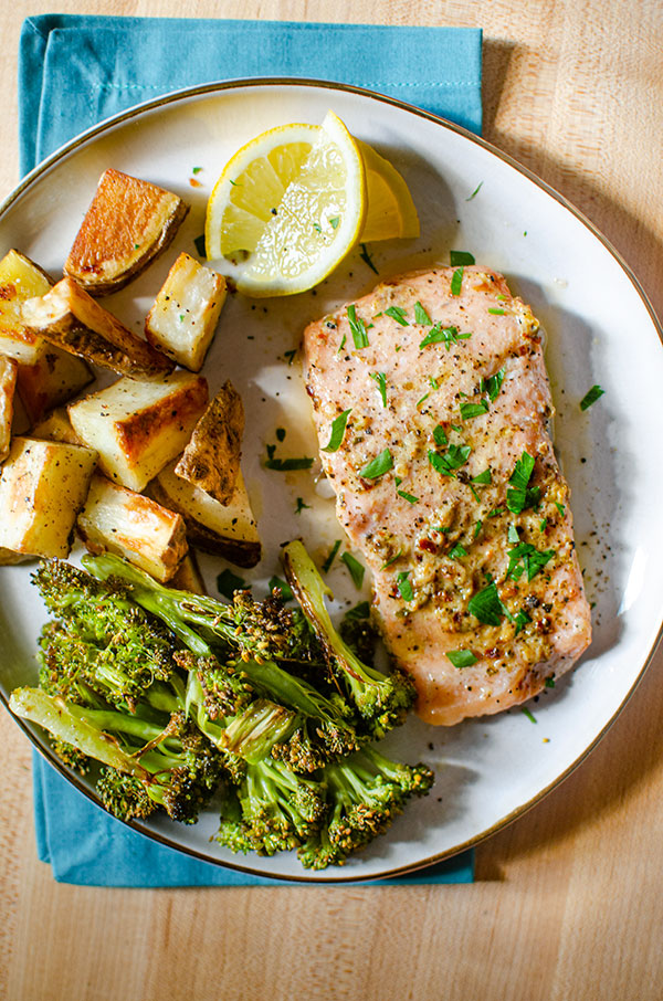 Baked trout fillet on a plate with roasted potatoes, broccoli and lemon wedges.