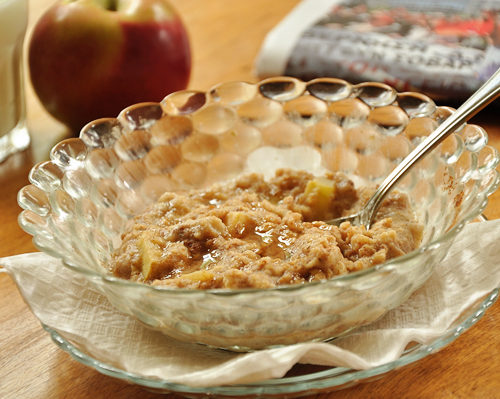 A bowl of apple cinnamon oatmeal with a glass of milk, an apple and a newspaper