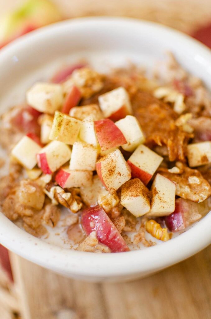 Closeup of a serving of oatmeal in a white bowl with chopped apples, peanut butter and walnuts on top.