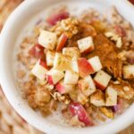 Closeup of apple cinnamon oatmeal in a white bowl with fresh chopped apples, peanut butter and walnuts on top.