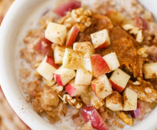 Closeup of apple cinnamon oatmeal in a white bowl with fresh chopped apples, peanut butter and walnuts on top.