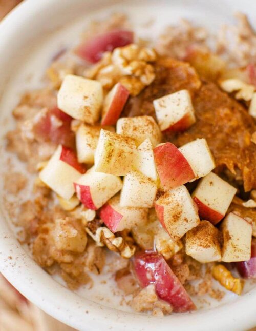Closeup of apple cinnamon oatmeal in a white bowl with fresh chopped apples, peanut butter and walnuts on top.