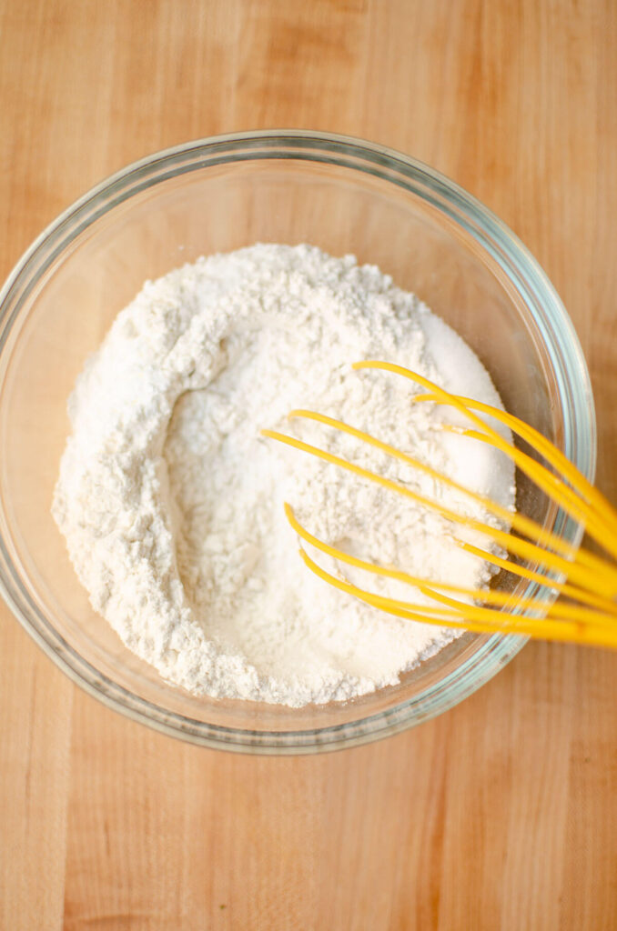 Dry ingredients in a glass bowl with a yellow whisk.