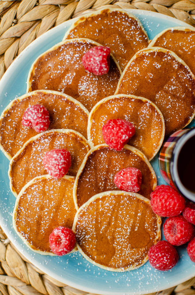 A blue platter of mini pancakes with raspberries scattered over top, with powdered sugar and maple syrup.