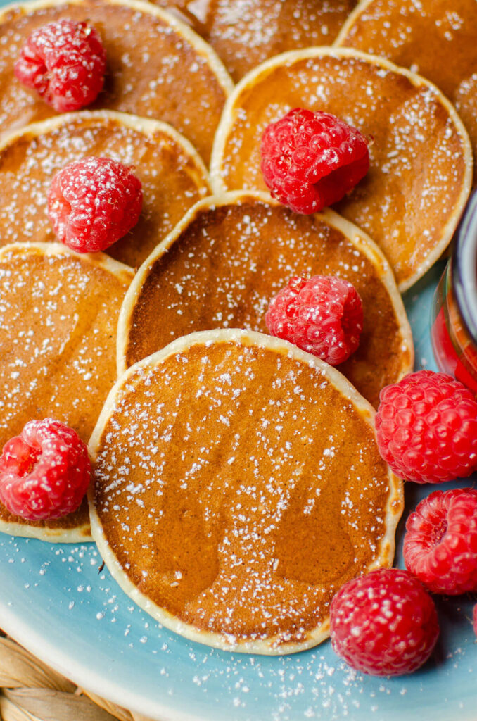 Closeup of mini pancakes on a blue plate with raspberries and maple syrup on top.