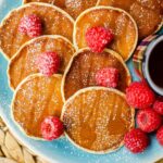 Six pancakes on a plate with raspberries sprinkled over top, with icing sugar and a small cup of maple syrup.