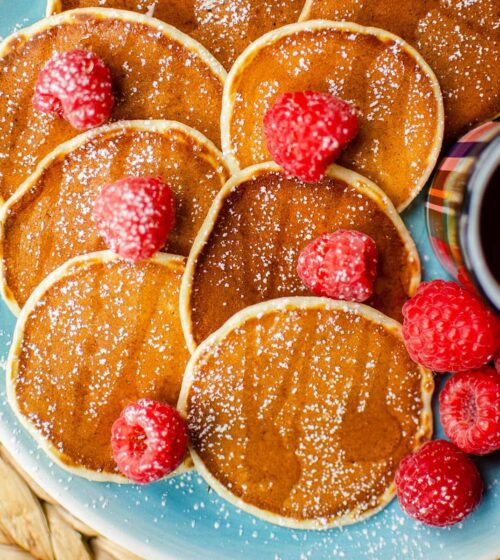 Six pancakes on a plate with raspberries sprinkled over top, with icing sugar and a small cup of maple syrup.