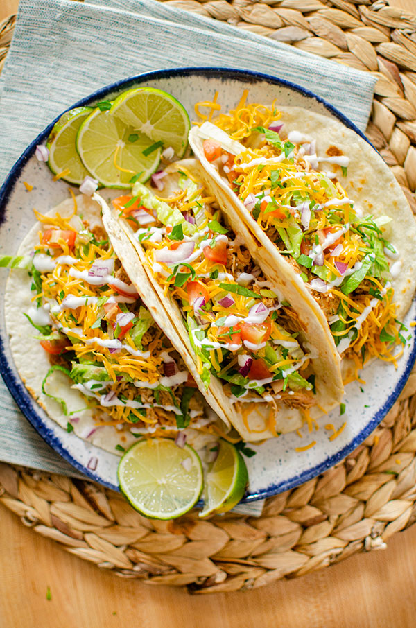 Three chicken tacos on a plate with all of the toppings; lettuce, tomatoes, cheese and sour cream.