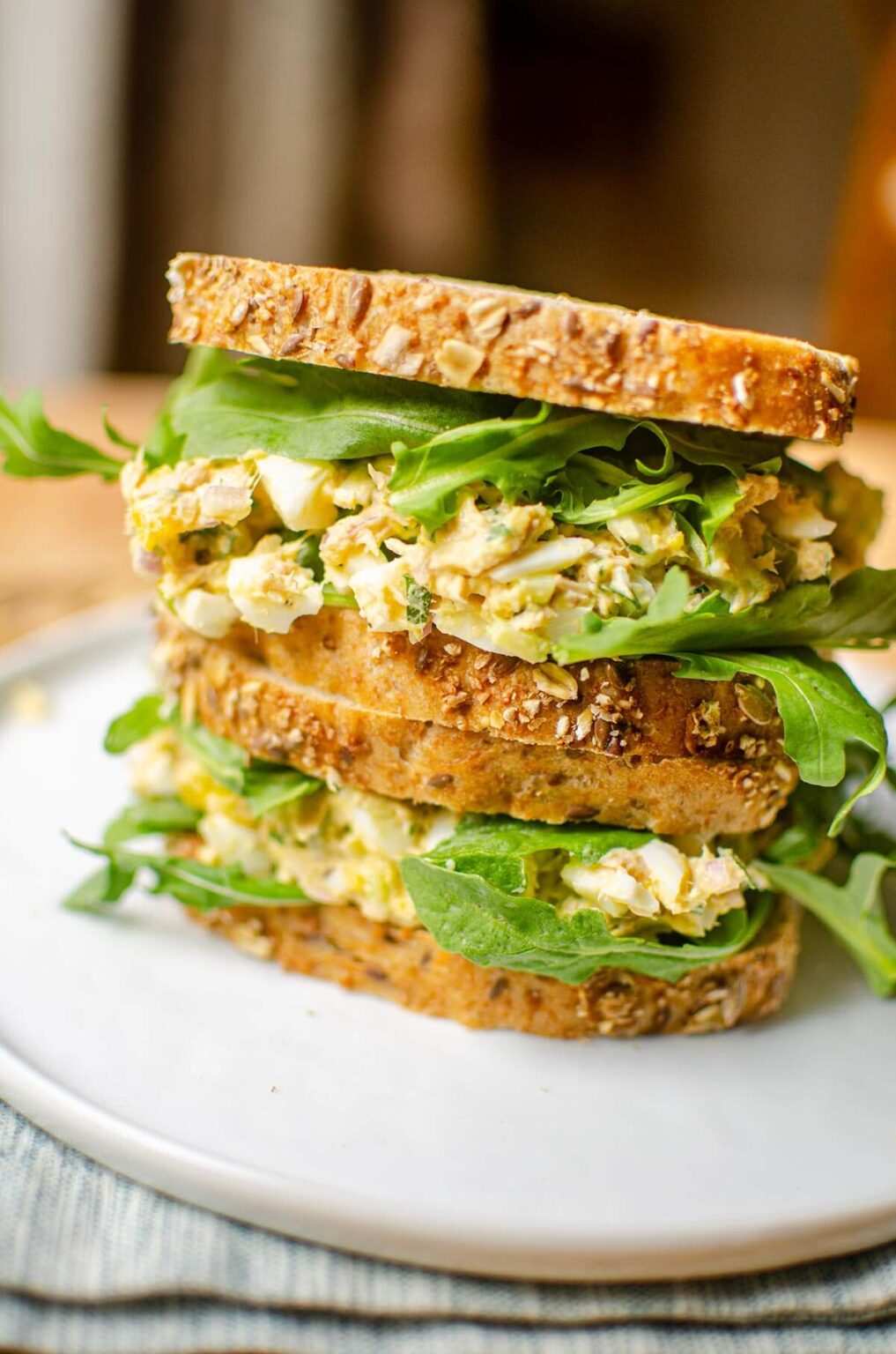 Two sandwiches stacked on top of each other filled with tuna egg salad and arugula on seeded whole grain bread.