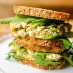 Two sandwiches stacked on top of each other filled with tuna egg salad and arugula on seeded whole grain bread.