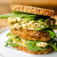 Two sandwiches stacked on top of each other filled with tuna egg salad and arugula on seeded whole grain bread.
