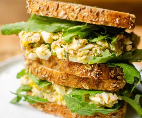 Two sandwiches stacked on top of each other filled with tuna egg salad and arugula on seeded whole grain bread.