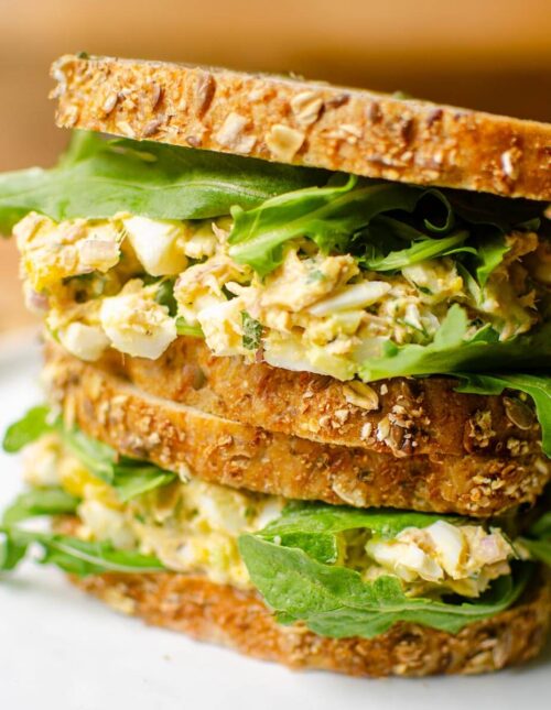 Two sandwiches stacked on top of each other filled with tuna egg salad and arugula on seeded whole grain bread.