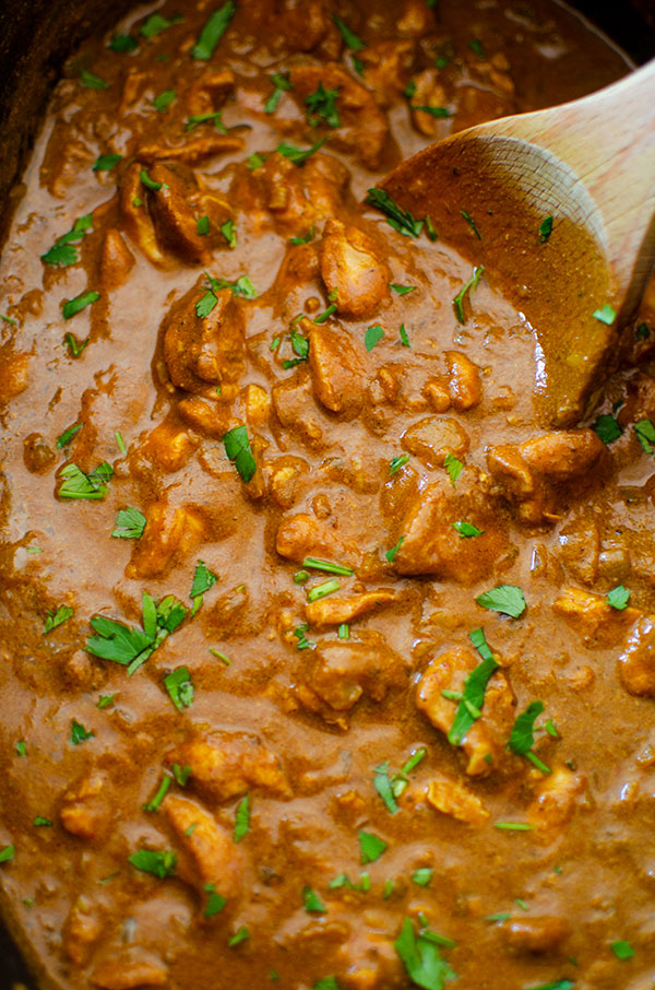 Slow cooker butter chicken in the slow cooker insert being stirred by a wooden spoon