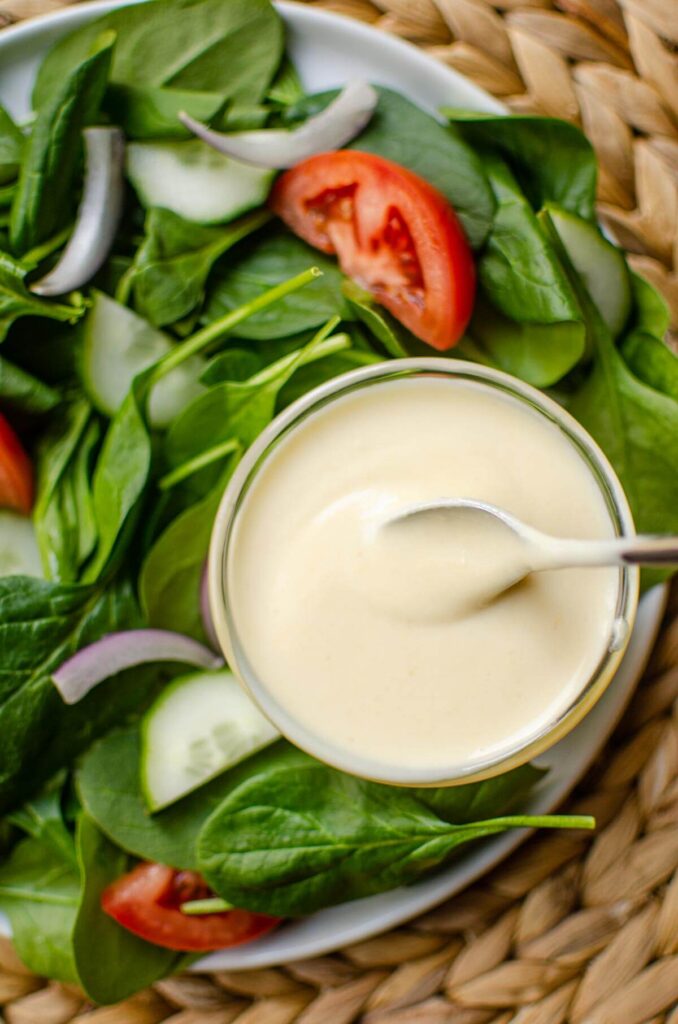 A jar of hummus dressing with a spoon on a plate with salad on the side.