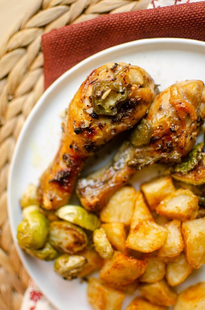 Two chicken drumsticks on a white plate with potatoes and Brussels spouts.