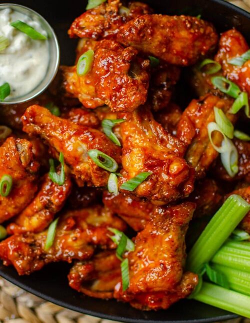 Closeup of chicken wings in a cast iron platter with celery and dip, with sliced green onions sprinkled on top.
