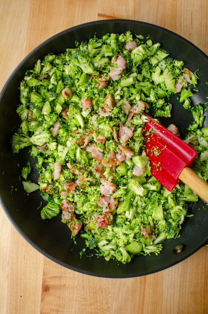 Bacon and broccoli in a pan being stirred by a red spatula.
