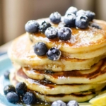 Closeup of a stack of pancakes with fresh bluberries, maple syrup and icing sugar on a blue plate