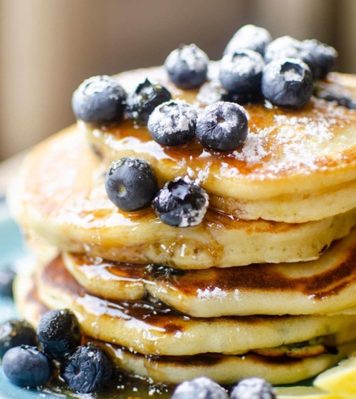 Closeup of a stack of pancakes with fresh bluberries, maple syrup and icing sugar on a blue plate