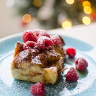 A slice of eggnog French toast with raspberries and powdered sugar with a Christmas tree in the background.