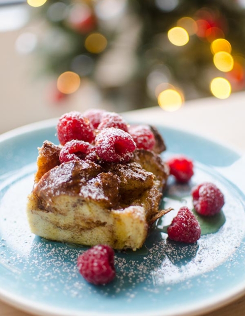 A slice of eggnog French toast with raspberries and powdered sugar with a Christmas tree in the background.
