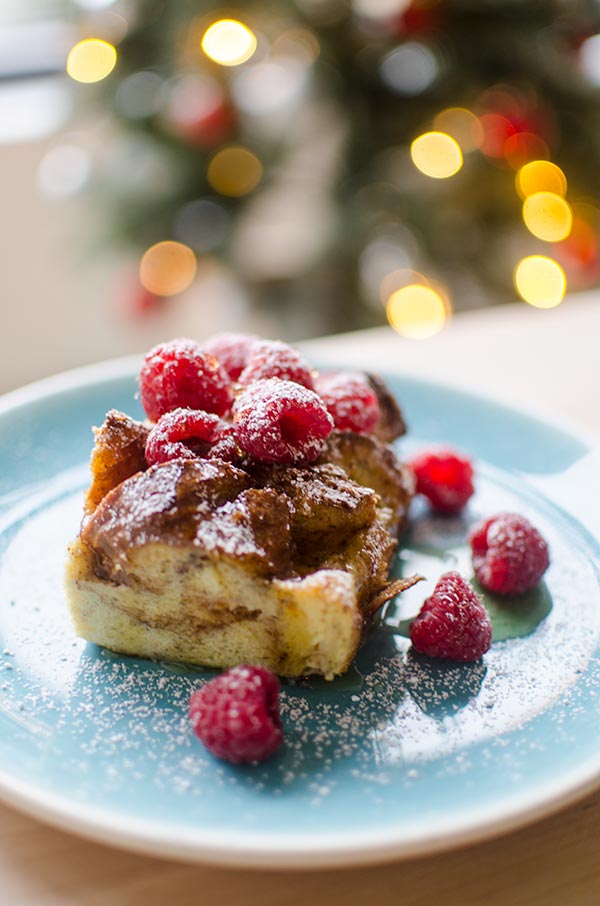 A slice of eggnog French toast with raspberries and powdered sugar with a Christmas tree in the background.