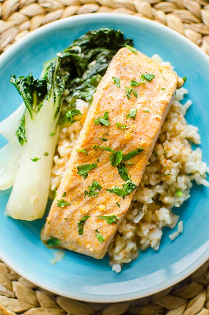 Fillet of salmon overtop brown rice with a side of bok choy on a turquoise plate.