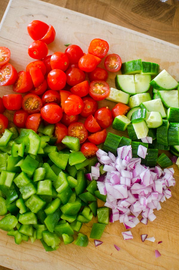 Ingredients (cherry tomatoes, cucumber, green pepper and red onion) prepped on a cutting board.