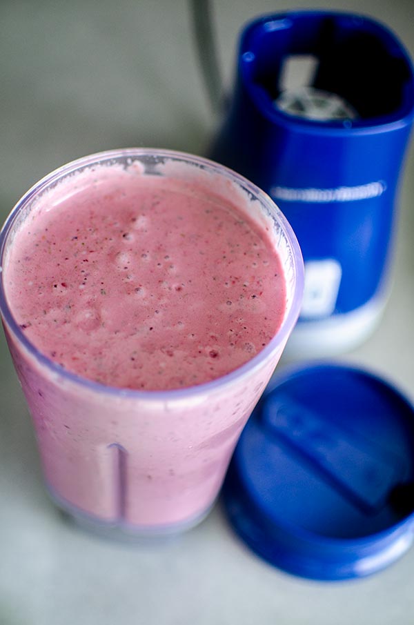 Closeup of open smoothie container with red triple berry smoothie next to blender.