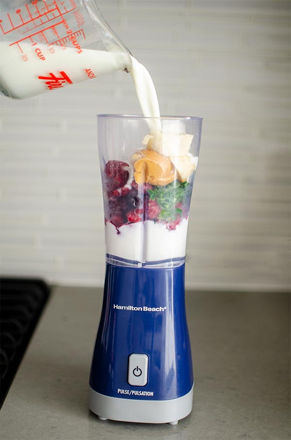 Milk getting poured into open blender with yogurt, berry mix, peanut butter and spinach.