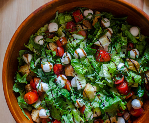 A bowl of bruschetta salad with balsamic drizzle on a wooden board.