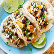 Three cauliflower tacos on a blue plate with sliced limes and a crema drizzle