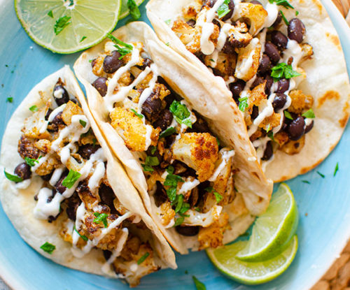 Three cauliflower tacos on a blue plate with sliced limes and a crema drizzle