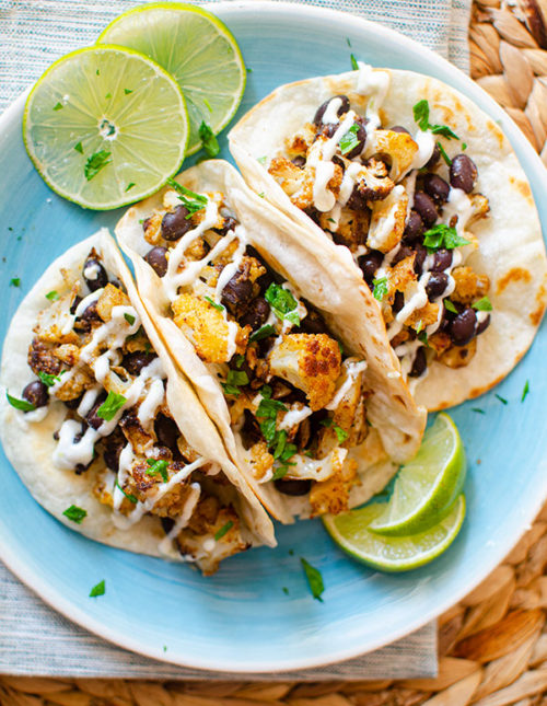 Three cauliflower tacos on a blue plate with sliced limes and a crema drizzle