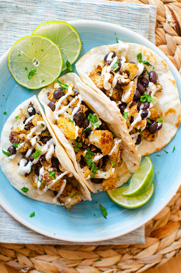 Three cauliflower tacos on a blue plate with sliced limes and a crema drizzle