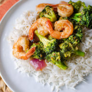 Shrimp and broccoli stir fry over rice on a white plate with an orange napkin