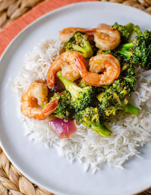 Shrimp and broccoli stir fry over rice on a white plate with an orange napkin