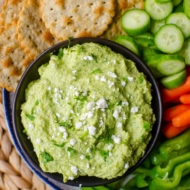 A bowl of dip with crackers, cucumber, red and green bell pepper