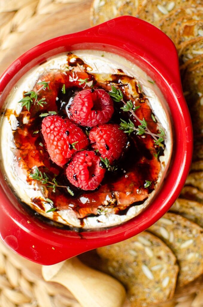 Baked brie in a red brie baker with crackers, topped with fresh raspberries.