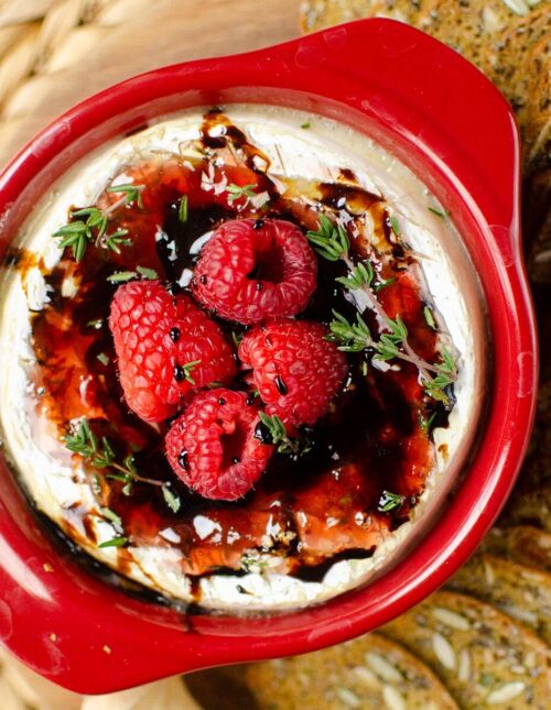Baked brie in red brie baker with raspberries and balsamic glaze on top.