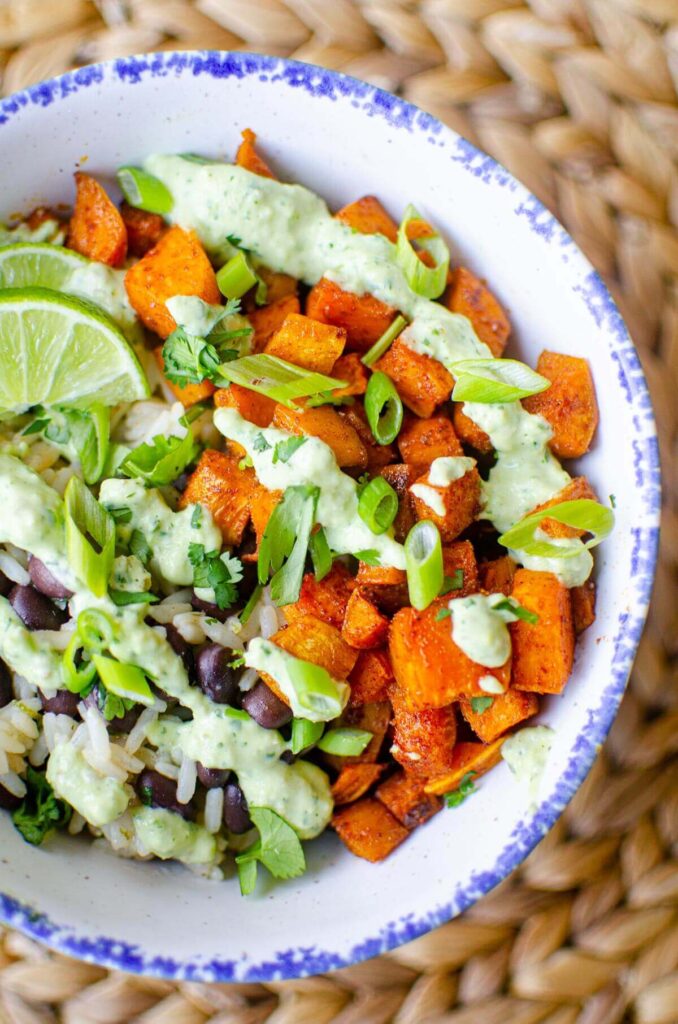 A serving of roasted sweet potatoes, black beans and rice with cilantro sauce drizzled overtop and sprinkled with green onions in a white bowl with a blue border.