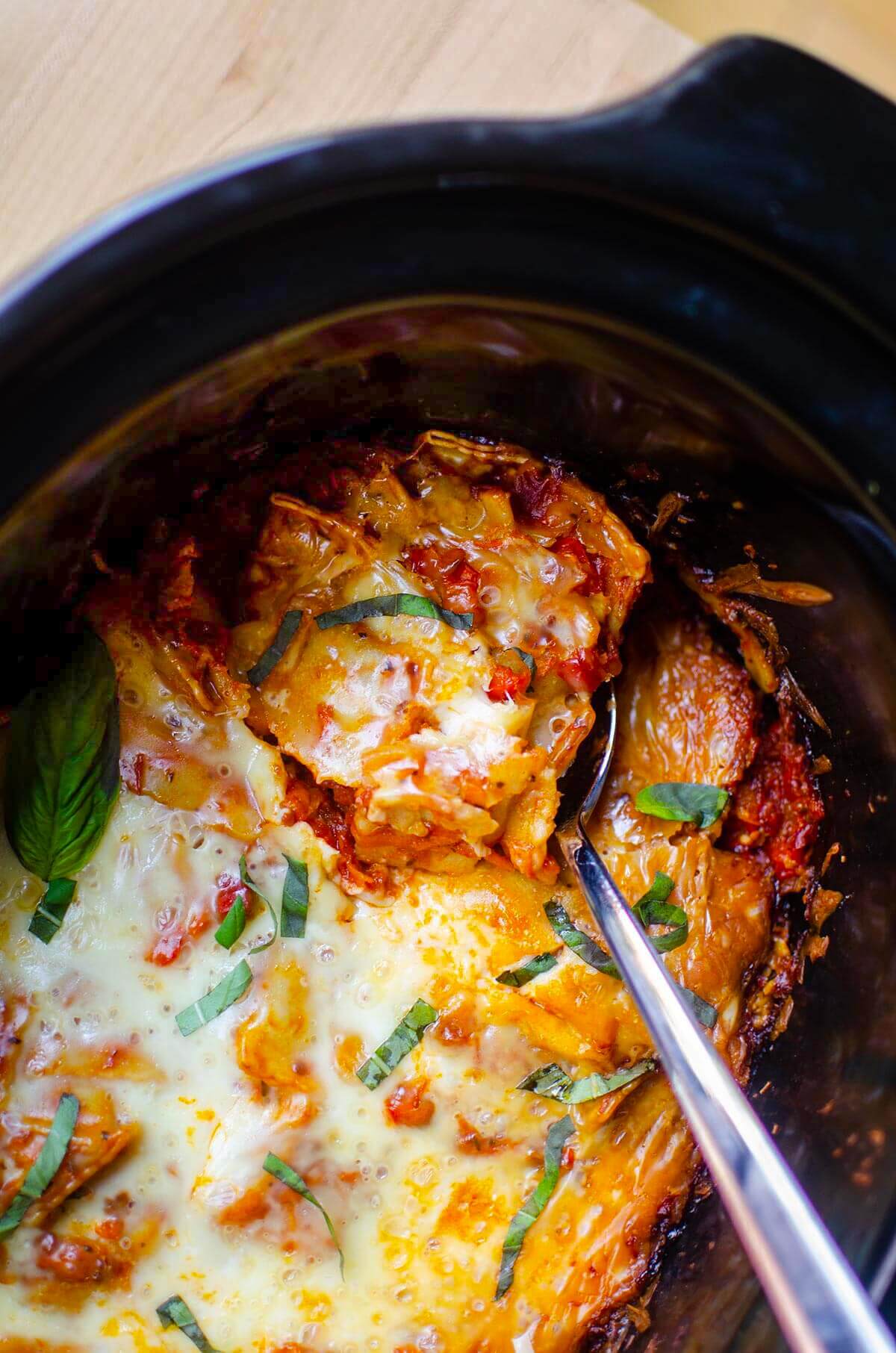 Ravioli lasagna in a slow cooker, with a spoon reaching in to grab a serving. Basil sprinkled overtop of melted cheese.