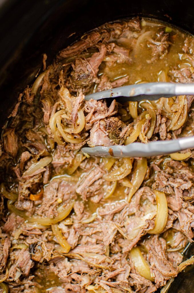 Closeup of shredded beef in the slow cooker being picked up by a pair of tongs.