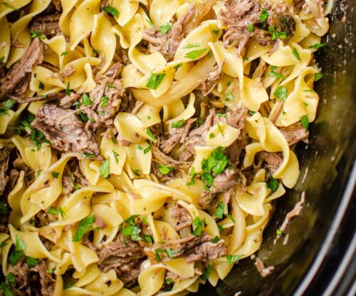 Closeup of pulled beef in the slow cooker with egg noodles with parsley sprinkled over top.