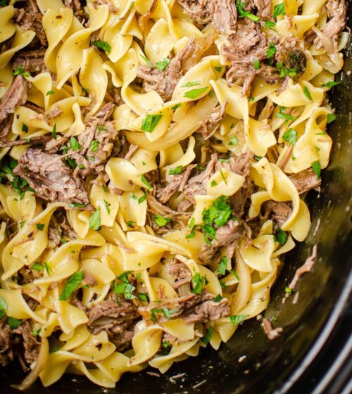 Closeup of pulled beef in the slow cooker with egg noodles with parsley sprinkled over top.